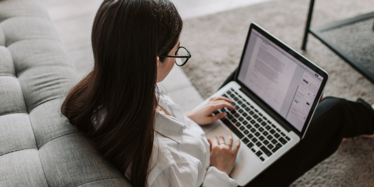 Kvinna sitter i en soffa med en laptop i knät och läser ett dokument.