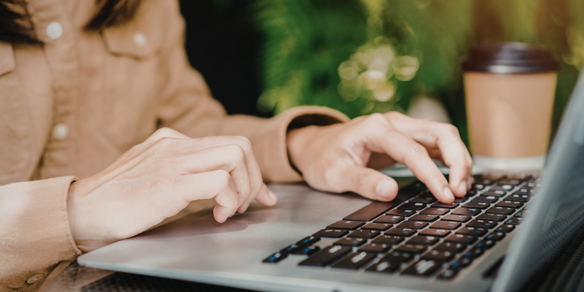 Person skriver på en bärbar dator vid ett bord, med en take away-kaffe bredvid och grönskande bakgrund.